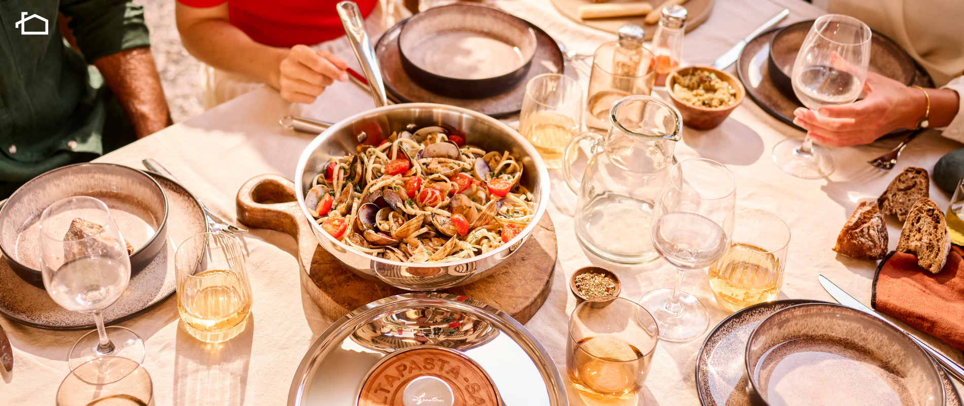 A stainless steel Saltapasta pan filled with spaghetti, clams, and cherry tomatoes sits at the center of a sunlit outdoor dining table, surrounded by glassware, plates, bread, and people enjoying a meal.